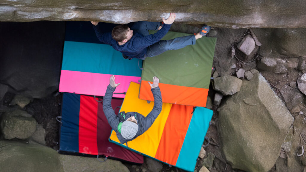 boulderer climbing with 5 colorful crashpads below and another person spotting